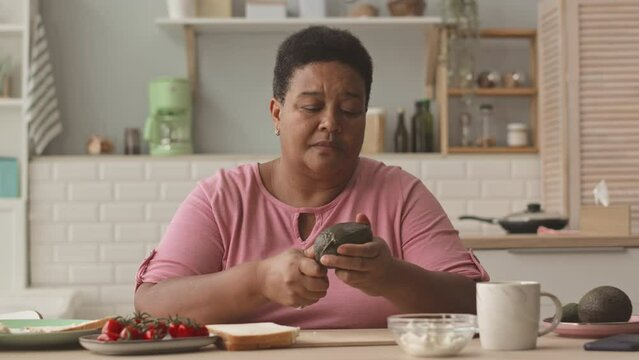 Waist up of mid aged African American woman cutting avocado while making toasts for breakfast at bright cozy kitchen