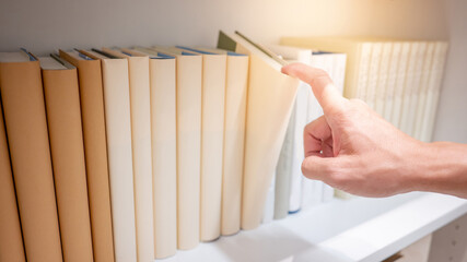 Male hand choosing and picking light beige book on wooden bookshelf in public library. Education...