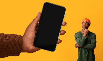Thoughtful Young Black Guy Looking At Huge Blank Smartphone In Big Hand