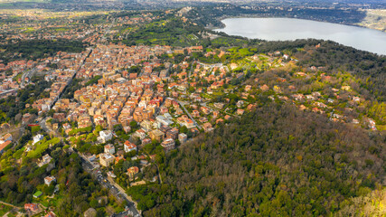 Fototapeta na wymiar Aerial view of Lake Albano, a volcanic crater lake. A luxuriant forest grows around the water and on the left there is the city of Albano Laziale, in the Metropolitan City of Rome, Italy.