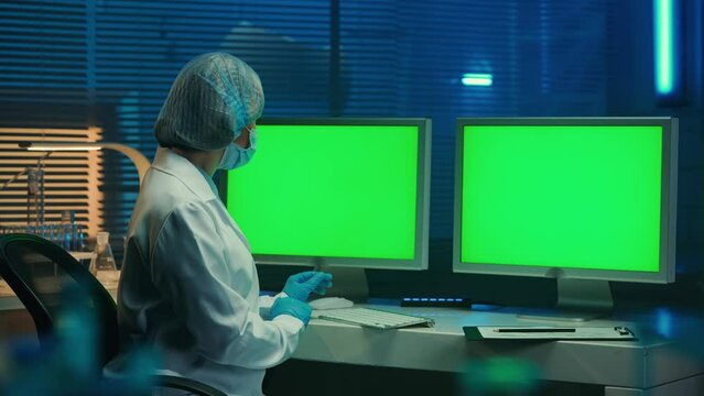 A doctor typing on a computer keyboard with green screen. Template place for your text or image, promotional content. Advertising area, workspace mock up. A woman in a white gown, gloves and bonnet.