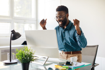 Overjoyed black businessman celebrating success with computer, looking at monitor and raising fists...
