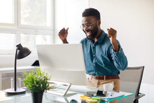 Overjoyed black businessman celebrating success with computer, looking at monitor and raising fists with excitement