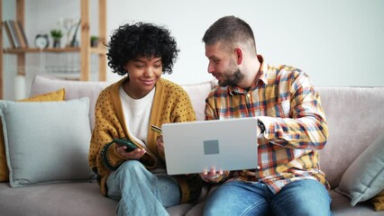 Positive international couple shopping online together, african woman with a debit card and caucasian man with the laptop making an order, purchase online. Happy multiracial couple shopping online. - Powered by Adobe