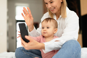 Smiling mother and her little daughter having video call on smartphone, sitting on bed and waving at phone screen