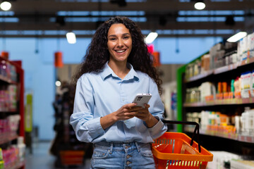 Shopping in a supermarket. A young Latin American woman stands between rows of shelves with a cart....