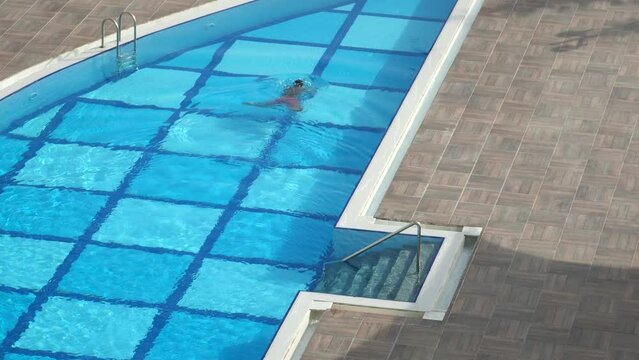 Top down aerial view of young man swims in a swimming pool outdoor in breaststroke technique. Man relaxes at opened swimming pool. High angle view