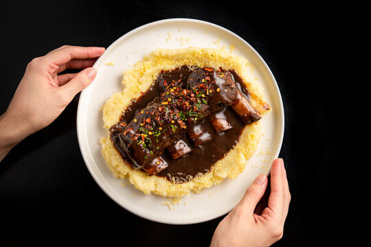 plano cenital de unas costillas de res con picante sobre un pure de papas con queso y unas manos de mujer sosteniendo el plato en un fondo negro