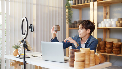 Asian young adult man is giving a presentation about vase product by live streaming online at home. Active male merchant is showing and explaining details of goods to online customers by smartphone.