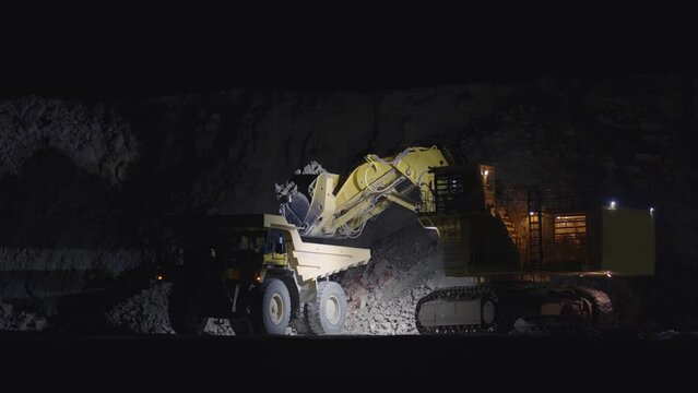 Large escavator loading ore rock in dumper truck at night, slow motion