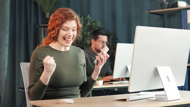 Happy red hair business woman in office looking at mobile phone with emotion winner or win, financial stock sports betting. Excited overjoyed girl celebrating success,makes hand gestures says yes.
