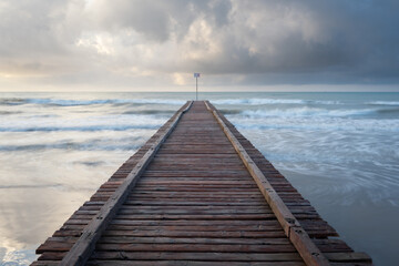 Fototapeta premium a wooden pier leading into a rough sea under a cloudy sky. long exposure