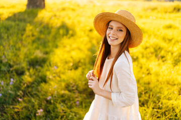 Portrait of lovely redhead young woman in straw hat and white dress standing posing on beautiful meadow of green grass looking at camera, on background of warm sunlight at summer day.