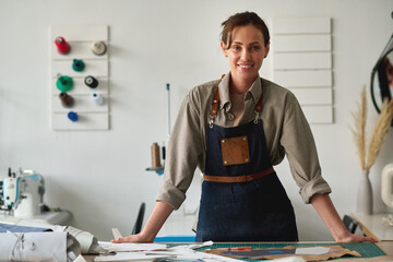 Young successful female fashion designer or tailor in workwear standing by her workplace in atelier...