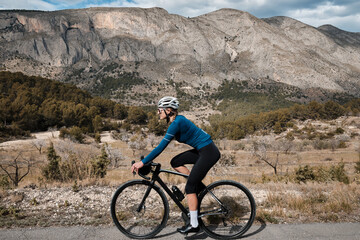 Obraz premium With a view of the beautiful mountains, a woman cyclist wearing a cycling kit and helmet is riding a gravel bike. Alicante region in Spain.