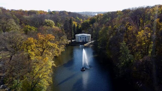 Beautiful landscaped park with lake, fountain, trees, large stones, people walking and white architecture on sunny autumn day. Trees with yellow leaves on shores, tourists high flowing jet of fountain