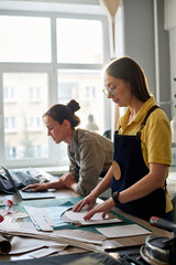 Fototapeta premium Side view of female tailors working in atelier or studio of fashion while one of them networking and the other going to circle sewing patterns