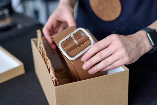 Hands of young female tanner putting new handmade folded leather belt of brown color into cardboard box while packing it for client