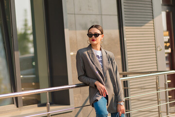 Incredible portrait of amazing girl on the street in sunlight. She wears jeans, grey jacket and dark glasses. Stylish woman with red lips. Fashion concept