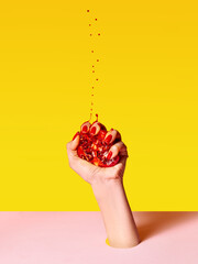 Human hand with red painted nails squeezing half of pomegranate over pink yellow background. Contemporary art. Drops of juice fly up
