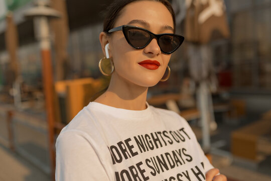Incredible pretty woman with red lips wearing dark sunglasses and stylish white t-shirt posing to camera in summer day. Portrait of European girl on sunset background