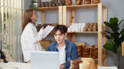 Asian young adult man is telling to Caucasian female coworker to send vase products to him for checking the online purchase order. Two male and female merchants are busy preparing for shipping goods.