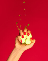 Closeup female hand with red painted nails squeezing half of apple fruit over magenta background. Contemporary art. Drops of juice fly up