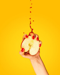 Closeup female hand with red painted nails squeezing half of apple fruit over yellow background. Contemporary art. Drops of juice fly up