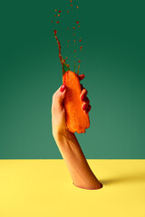 Elegant woman's hand squeezes carrot with force and drops of juice fly up over green yellow background. Pop art photography.
