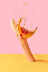 Female hand with bright manicure squeezes banana over pink-yellow background. Pop art food photography. Drops of juice fly up