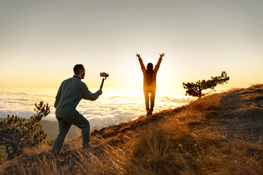 Man tourist with electronic stabilizer gimbal is taking video shoot with happy girl at sunrise mountain top