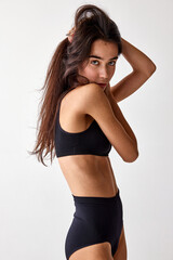Fototapeta premium Portrait of beautiful young brunette girl with slim fit body posing in black underwear over grey studio background. Concept of natural beauty, diet, femininity, wellness, body care and fitness