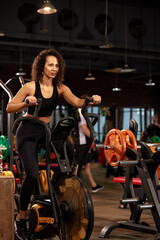 Fototapeta na wymiar Positive smiling african american woman, exercising on an exercise bike in the gym, dark background, cardio training on an exercise bike, keep yourself in good shape.