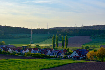 Rural houses with solar panels on the roofs. Plowed fields are in the foreground. Wind power plants in the background