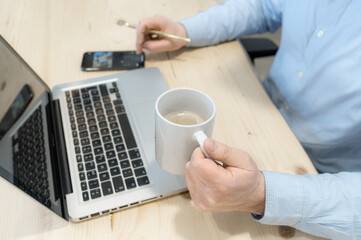 Arms and hands of a Caucasian man, working with a laptop, a mobile phone and having a cup of coffee, light wooden background