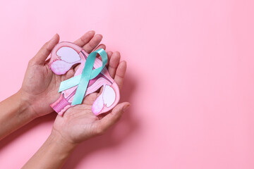 Teal awareness ribbon with cervix shape on palms hand over pink background with copy space. Ovarian Cancer month, cervical cancer day. 