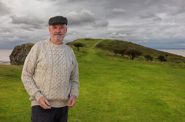 An elderly man wears a stylish Irish sheep's wool sweater and tweet cap. He on a green grassy area...