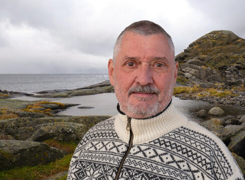 An elderly man with a beard is wearing a fashionable Norwegian sweater with a pattern typical of the country. The Norwegian coast and a mountain can be seen in the background.