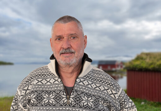 An elderly man with a beard is wearing a fashionable Norwegian sweater with a pattern typical of the country. The Norwegian coast and a hut can be seen in the background.