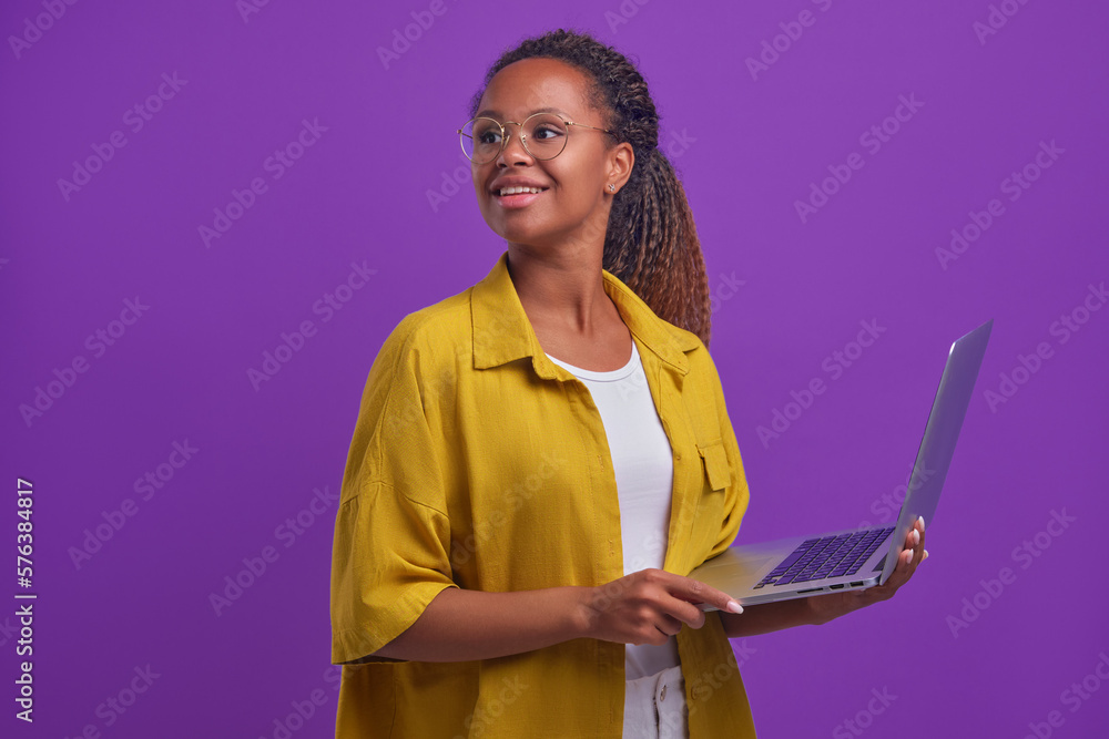 Wall mural Young beautiful happy African American woman with open laptop look back hearing voice of colleague doing remote work or getting online education stands on plain lilac background. Freelance, internet