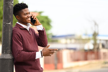 Young African man making a smartphone call and smiling boldly on the street .Handsome and excited young african wearing a half zip sweater on sweater. © The Yudel Media