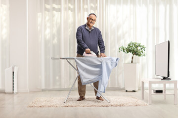 Mature man ironing a shirt and smiling in front of tv