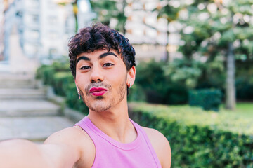Content young Hispanic male in pink tank top with makeup looking at camera while taking self portrait on street against green trees