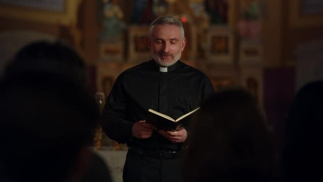 A priest in a black robe stands in front of the people during the Sunday service. The priest reads a passage from the Bible to the parishioners.