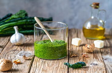 Homemade kale pesto served in a glass jar on wooden surface with grey background and basic...