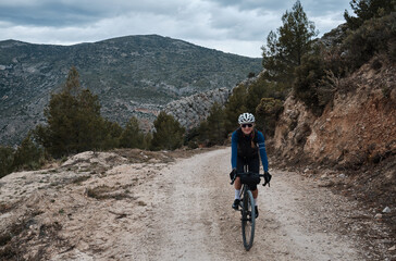 A woman cyclist is riding against a mountainous background.Female cyclist is riding a gravel bike on a gravel road.Cycling adventure. Parcent, Alicante, Spain