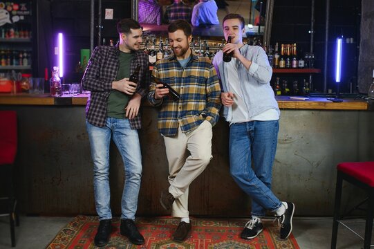 Three young men in casual clothes are smiling, holding bottles of beer while standing near bar counter in pub