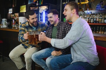 Three young men in casual clothes are smiling and clanging glasses of beer together while sitting at bar counter in pub