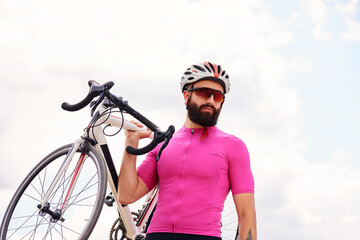 Bearded male cyclist in glasses and a helmet looks at the camera with a bicycle in his hand against...
