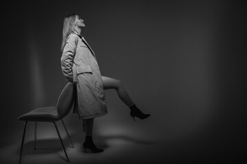 space, young, beautiful, emotional girl in a dark raincoat stands near a chair, raised one leg, leaning on a white background, a ray of light. Low key, ray of light
